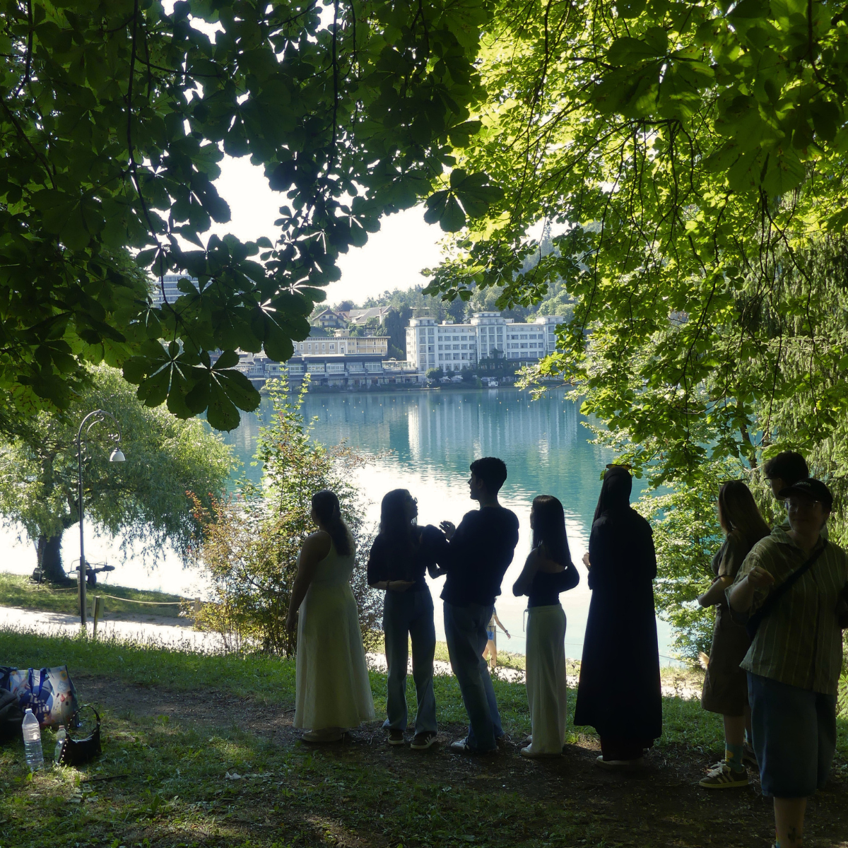 Gruppe junger Menschen steht im Schatten unter Bäumen und blickt auf den See